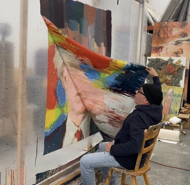Artist Bradley Hart sits on a wooden chair in his studio, lifting a colorful, paint-saturated sheet from a large bubble wrap surface to reveal the photorealistic portrait underneath. The artwork is made using acrylic paint injected into individual bubbles.